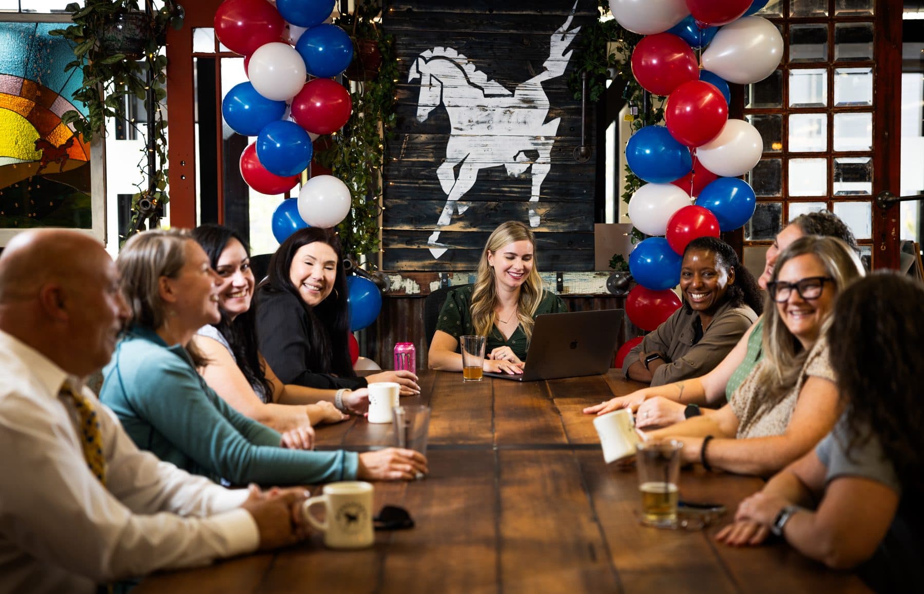 Members seated along the long table for a community lunch, balloons and a world map behind them.