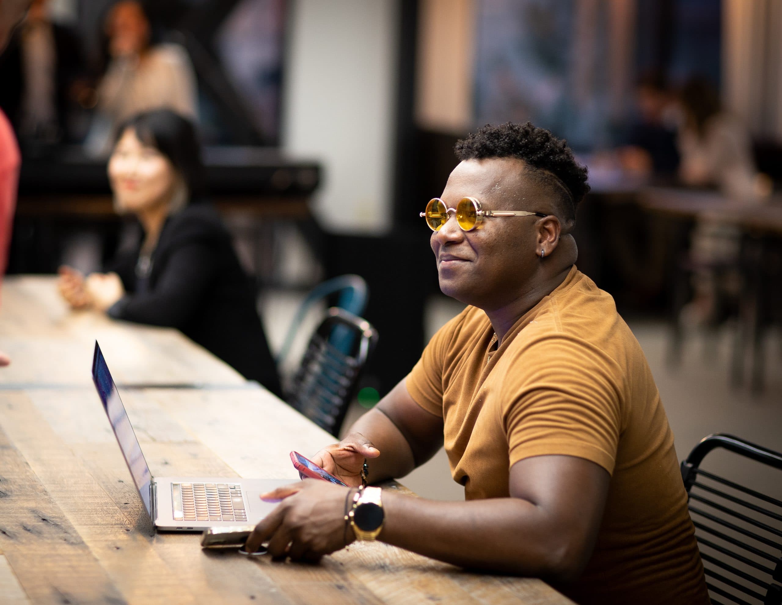 A member in a mustard shirt looks up from his laptop at the long communal table, with others working behind him.