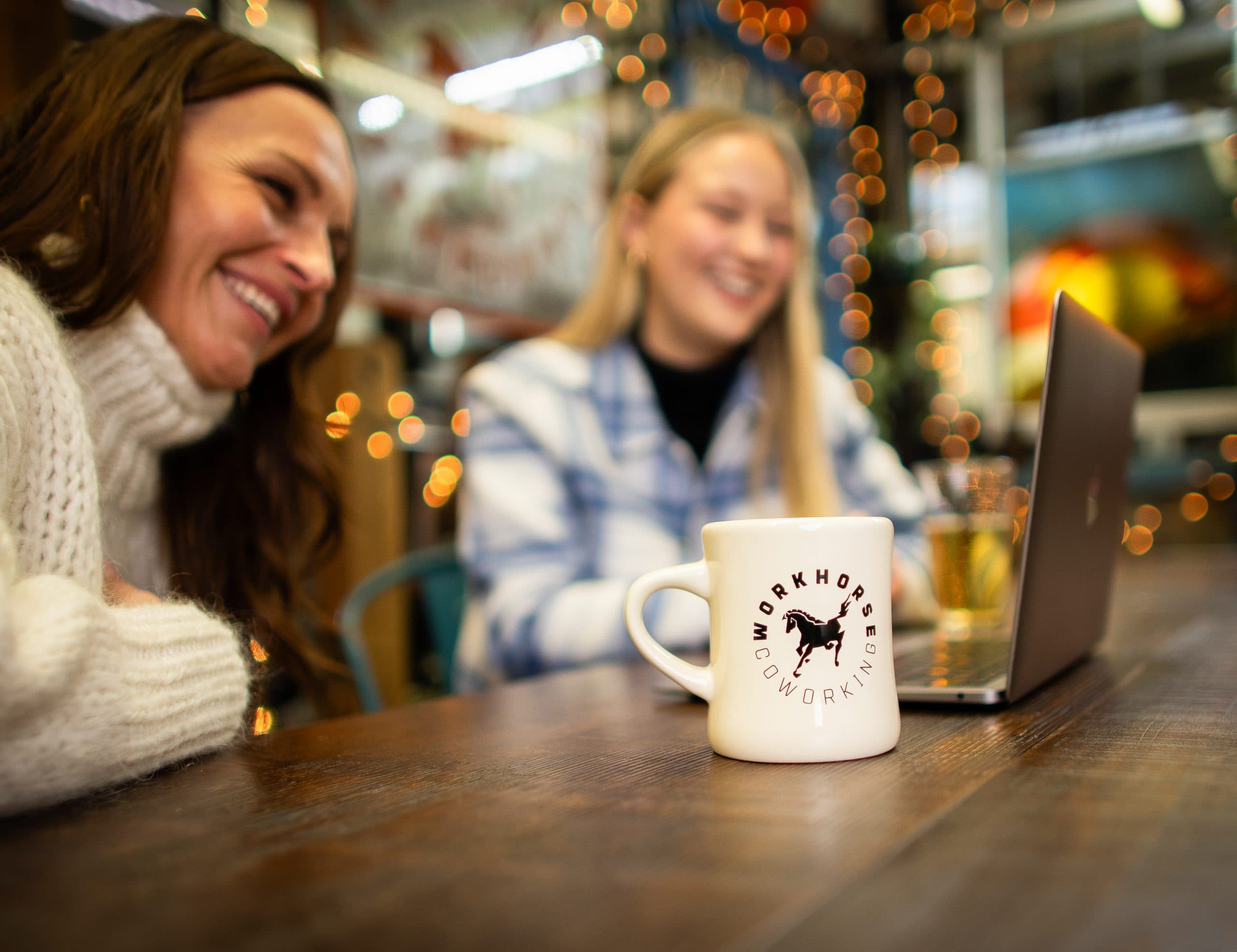Two members laughing together over a laptop, a branded Workhorse mug in the foreground.